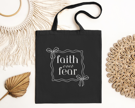 Christian tote bag with text "Faith Over Fear" and simple bow graphic. Cotton canvas bag for daily use. 